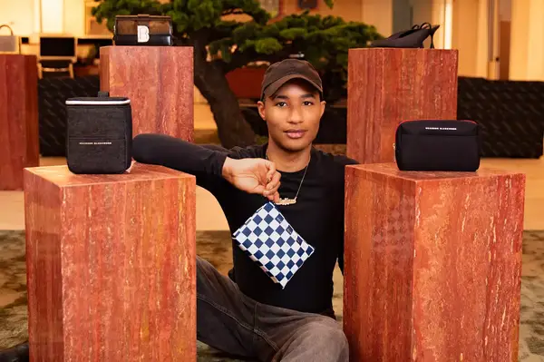 An individual seated among display pedestals holding a checkered pouch, alongside Brandon Blackwood branded items