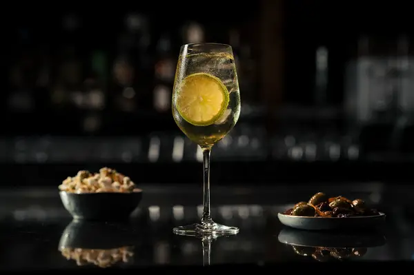 A drink with lemon garnish sits on a bar counter with small bowls of snacks