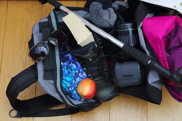 A packed hiking bag containing various items like a hiking pole, gloves, an apple, sunglasses, and a camera lens