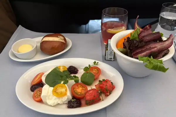 A meal served on an airplane includes a salad, bread roll, and a dish with burrata cheese and tomatoes