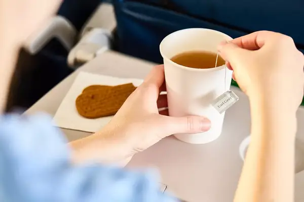A person holding a cup of tea with a biscuit on a tray table in an airplane