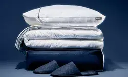 Stacked bedding items including pillows, folded linens, and slippers on a dark background