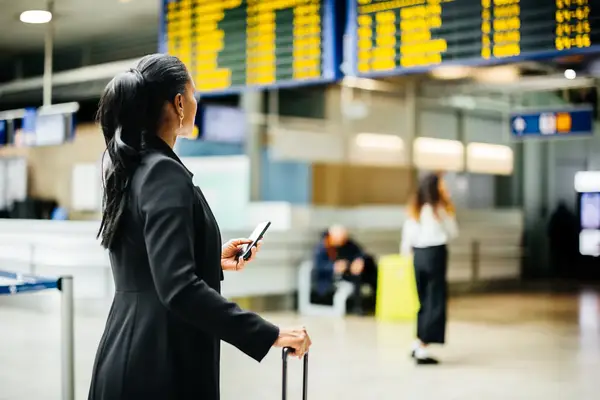 A woman standing in an airport terminal, holding a phone and suitcase, looking at the flight information board