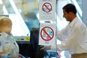 People at an airport security screening checkpoint signs showing prohibited items on the glass