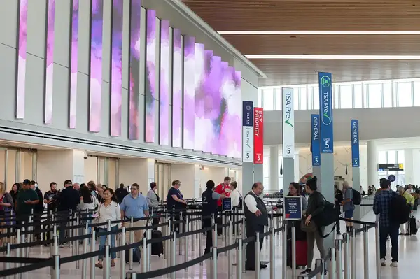 People standing in the TSA security line at an airport terminal with digital displays on the wall