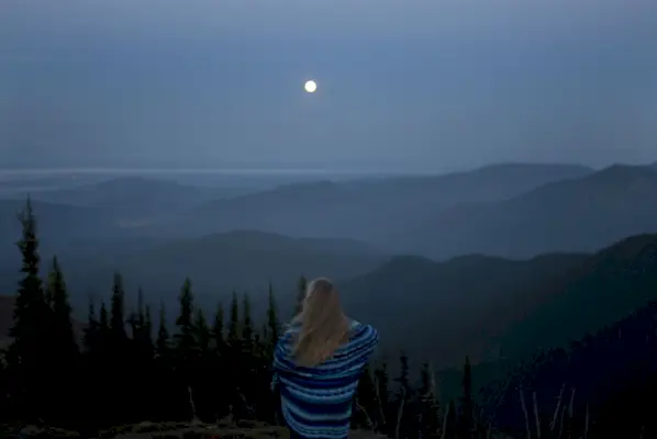 woman wrapped in blanket looking at mountainous landscape with full moon, rear view