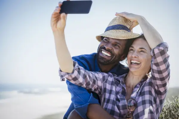 Playful, smiling multi-ethnic couple taking selfie with camera phone on sunny summer beach