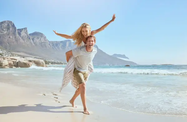 portrait of a happy and loving young couple enjoying a day at beach in summer cheerful affectionate husband giving his joyful wife a piggyback while walking and having fun by the seashore on holiday