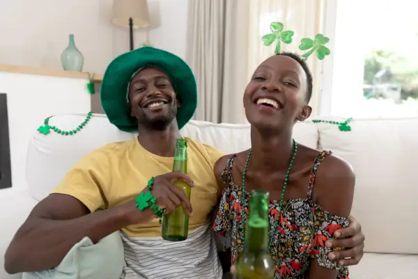 portrait of laughing african american couple in st patricks day costumes holding bottles of beer staying at home in isolation during quarantine lockdown