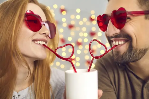 close up portrait happy young man and woman in love in pink sunglasses sipping drink from one cup through heart shaped straws, enjoying cute funny couple moment on a fun date on saint valentines day