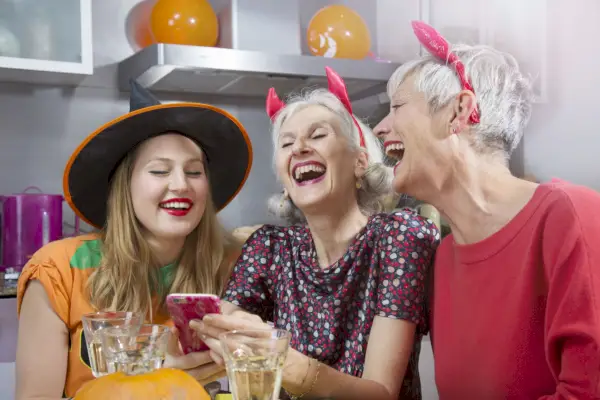 two older women in devil horns with younger woman in witch hat sitting a table laughing at something on a phone