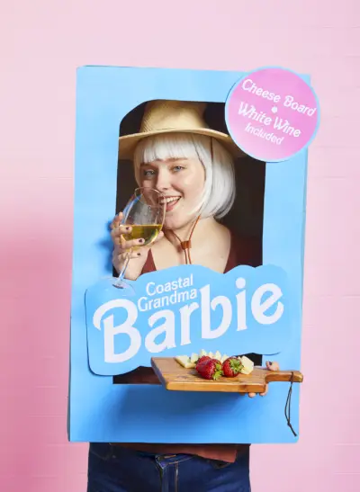 coastal grandma barbie costume featuring a woman in a blue cardboard barbie box holding a cheese plate and a glass of white wine