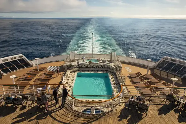 The deck of a cruise ship featuring a swimming pool with passengers overlooking the ocean at the ships wake