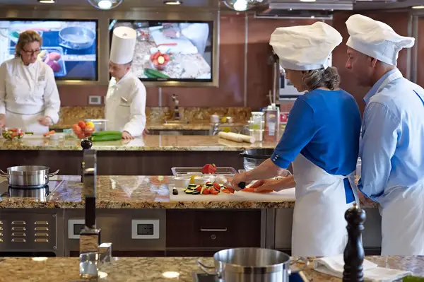 People cooking and preparing food in a culinary classroom, participants wearing chef hats at workstations