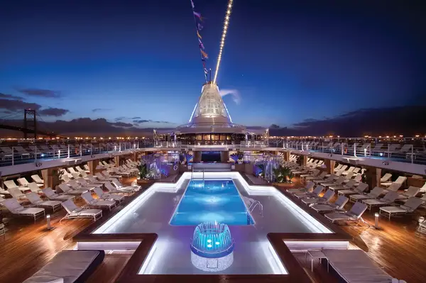 Pool deck of a cruise ship at night, featuring seating, a central illuminated pool, and deck chairs