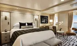 Luxurious bedroom interior in a ship suite, featuring a made bed with a headboard, seating area, and art on the wall