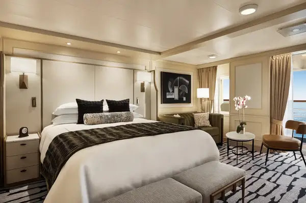 Luxurious bedroom interior in a ship suite, featuring a made bed with a headboard, seating area, and art on the wall