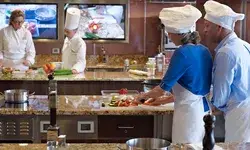 People cooking and preparing food in a culinary classroom, participants wearing chef hats at workstations