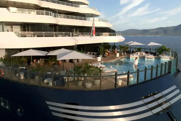 Luxury yacht stern featuring a pool area with sun loungers, umbrellas, and plants surrounded by glass railings, open water in the background