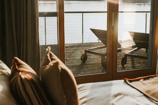 Serene, stylish interior bedroom with large sliding doors that lead out on to wooden decking. Two sun loungers are visible through the window. Vast lake can be viewed from the room and decking. Bed is covered with booster cushions. Sun shines through the window.