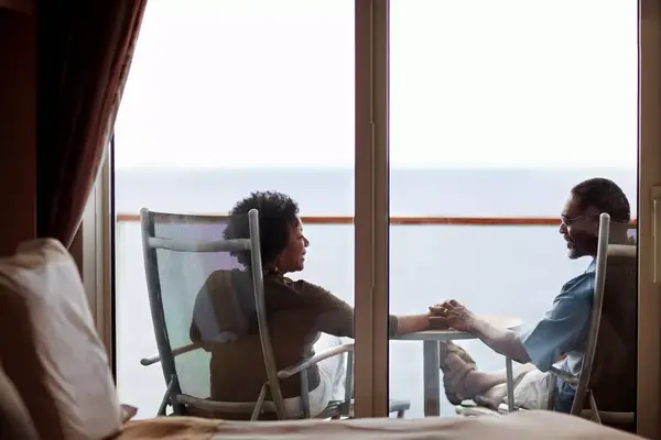 Mature couple sitting on chairs, holding hands, outside their cruise ship bedroom.
