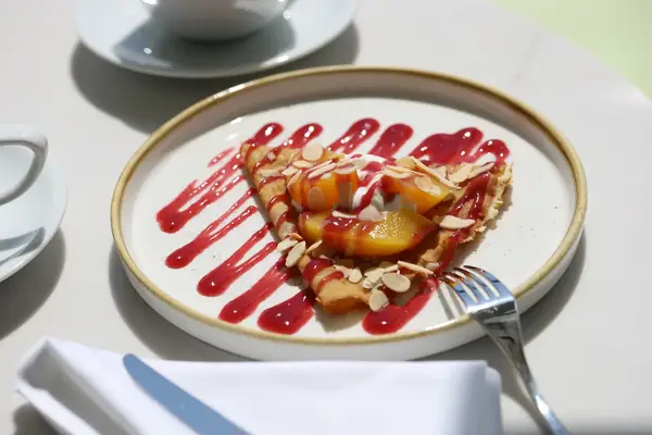 Plate with a crepe topped with peach slices almonds and red sauce drizzle served on a table with a fork and napkin