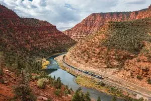 A train traveling along a river in a canyon with rocky treecovered hills