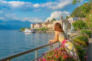 A person leaning on a railing next to a waterfront with buildings and mountains in the background