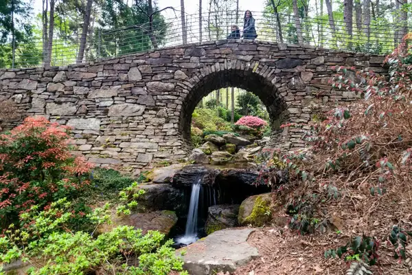A stone arch bridge over a small waterfall in a garden setting with a person sitting on top of the bridge