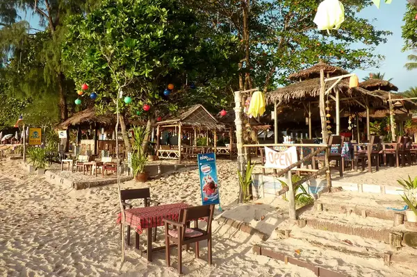 A beachside bar and restaurant with tables and chairs on the sand, surrounded by tropical trees and decorations