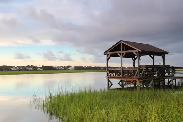 An empty wooden pavilion at the edge of a calm, grassy coastal area with buildings in the background