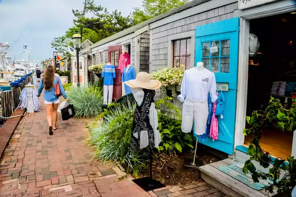 A cobblestoned shopping area with mannequins displayed outside stores, a person walking with a shopping bag in hand