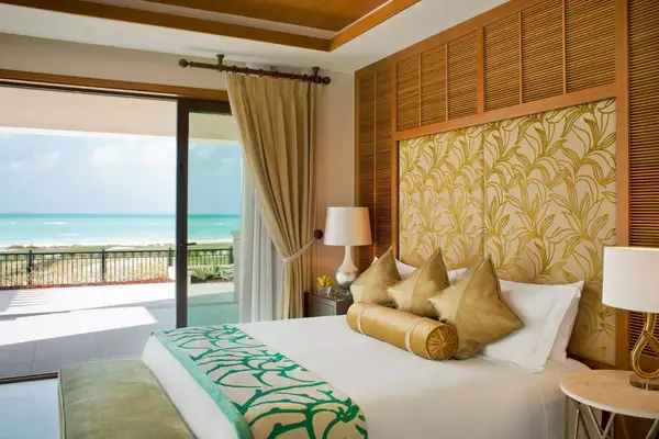 Luxury resort bedroom with balcony overlooking the ocean, bed with decorative pillows and patterned headboard, large sliding door nearby.