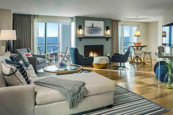 Living room of a hotel suite with ocean view through large windows, modern furniture including a fireplace, and a dining area