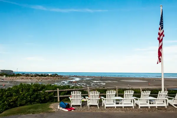 Adirondack chairs overlooking a sandy beach, ocean, and dunes, with a flagpole displaying the American flag