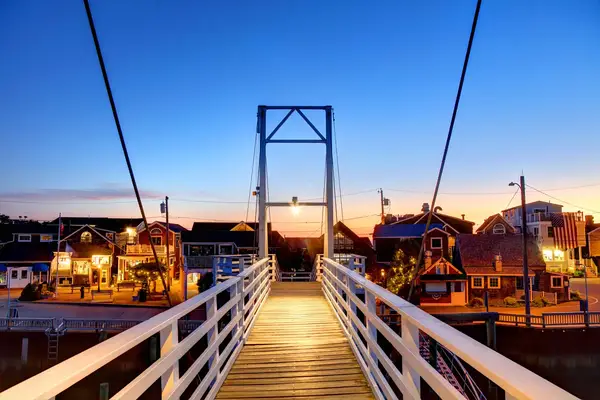 A pedestrian footbridge leading to a picturesque small town at twilight with lit buildings in the background