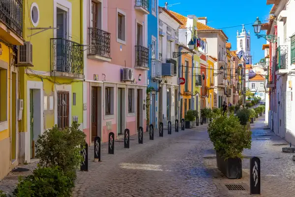 A cobblestone street lined with colorful buildings adorned with balconies plants along the street and a church tower in the background