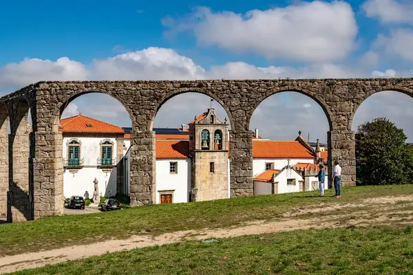 Stone aqueduct arches with buildings underneath and two people standing on grass in the foreground