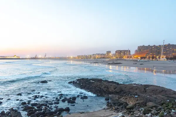 Coastal scene during sunset beach and rocky shore with buildings and lights in the background