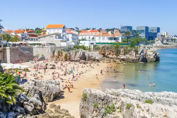 People enjoying a beach near a rocky coastline with buildings in the background