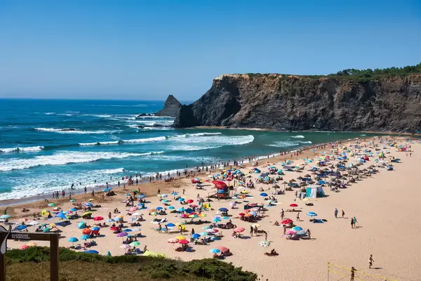 A crowded beach with sunbathers and umbrellas ocean waves and rocky cliffs in the background