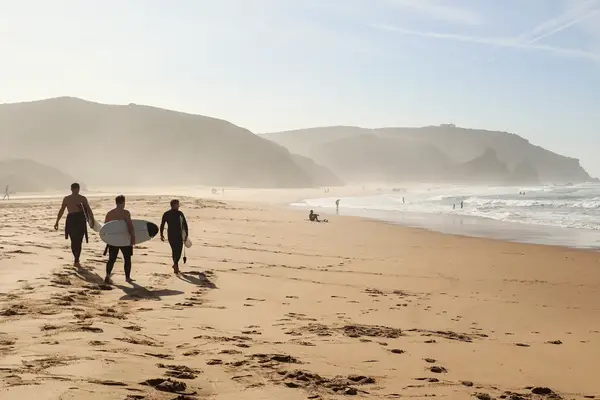 Three individuals walking on a sandy beach carrying surfboards waves and cliffs in the background