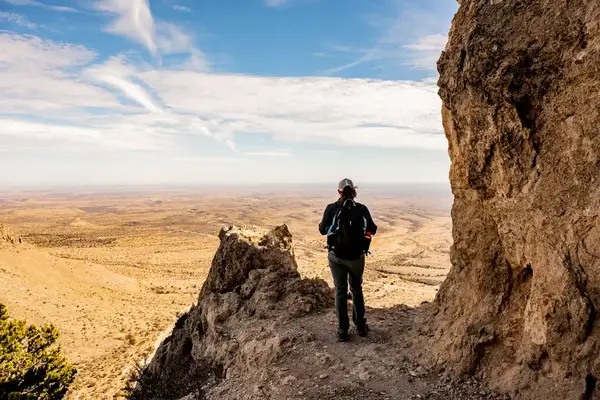 A hiker stands at a mountain viewpoint overlooking a vast desert landscape