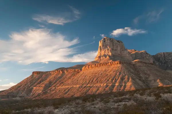 A desert mountain with striations and a prominent peak against a sky with scattered clouds