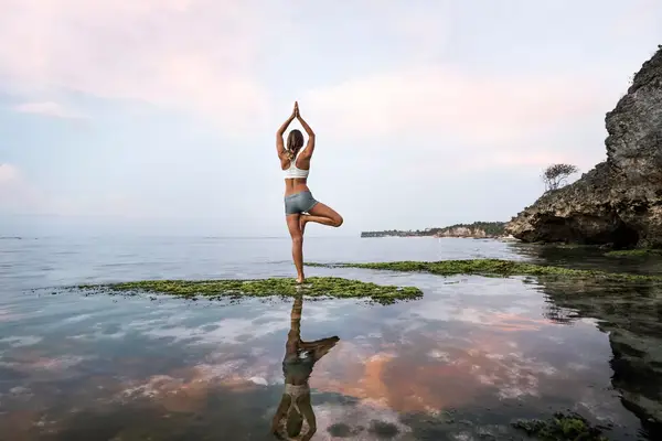 Asia, Indonesia, Bali, Uluwatu, young woman practicing yoga in front of rockpool at seashore, showing reflection in water