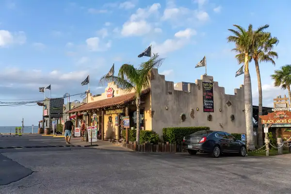 A tourist shop and boat tour office with pirate flags by the Laguna Madre in Port Isabel, Texas. Two older tourists stand in front.