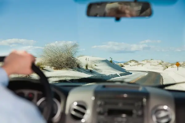 View from inside a car driving on a road through a desert area with sand dunes and sparse vegetation seen through the windshield
