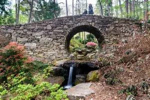 A stone arch bridge over a small waterfall in a garden setting with a person sitting on top of the bridge