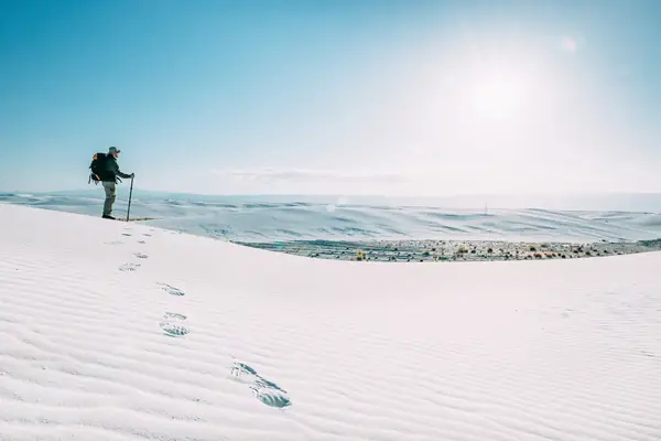 A hiker standing on sandy dunes with a backpack and walking stick overlooking a vast landscape