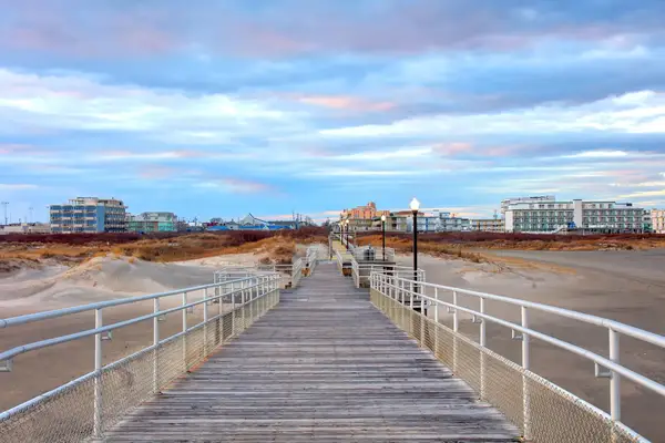 A wooden boardwalk extending over dunes towards buildings in the distance, a lamp post on the side